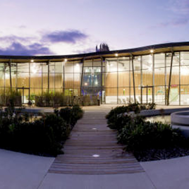 An aluminium and glass building is at the back of the image. It is very modern and angular. There are bushes and a pathway to the front of the image. 