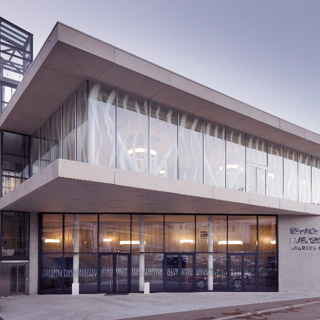 A glass and aluminium building. There is a window wall with black framing to the ground floor and large glass panels to the second floor.