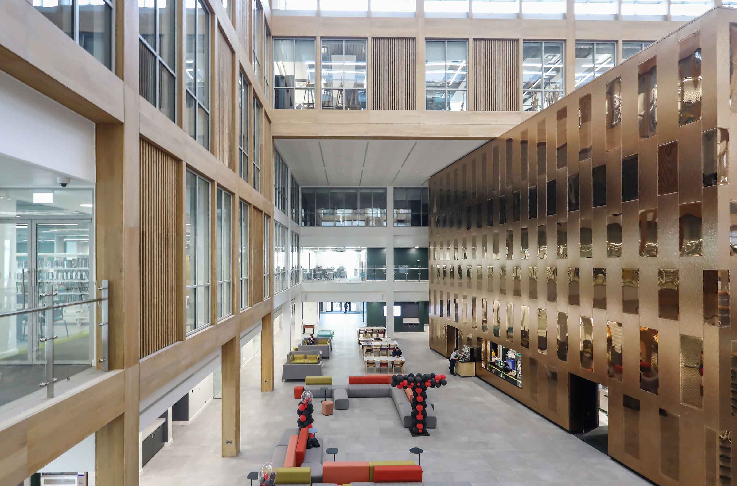 A corridor is shown. To the left, there is a glazed wall with gold coloured aluminium between the glass panes. To the right, there is a gold metal wall which is punctuated with windows. 