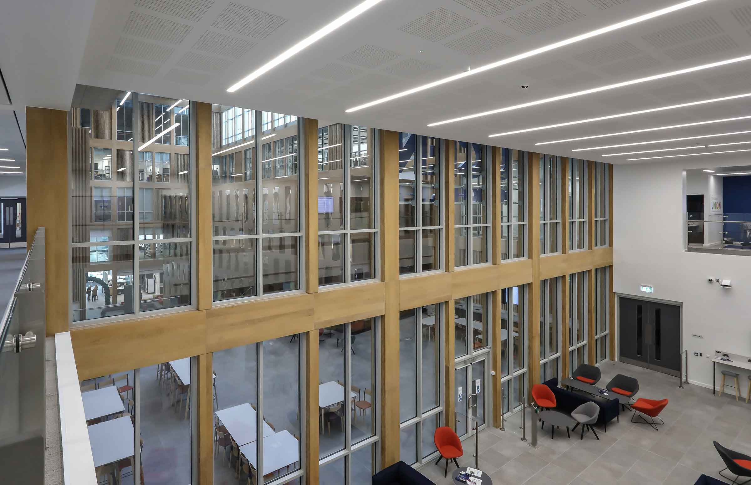 The inside of a modern educational building. There is a glass wall dividing the foyer and study space with chairs and tables for coffee on the floor.