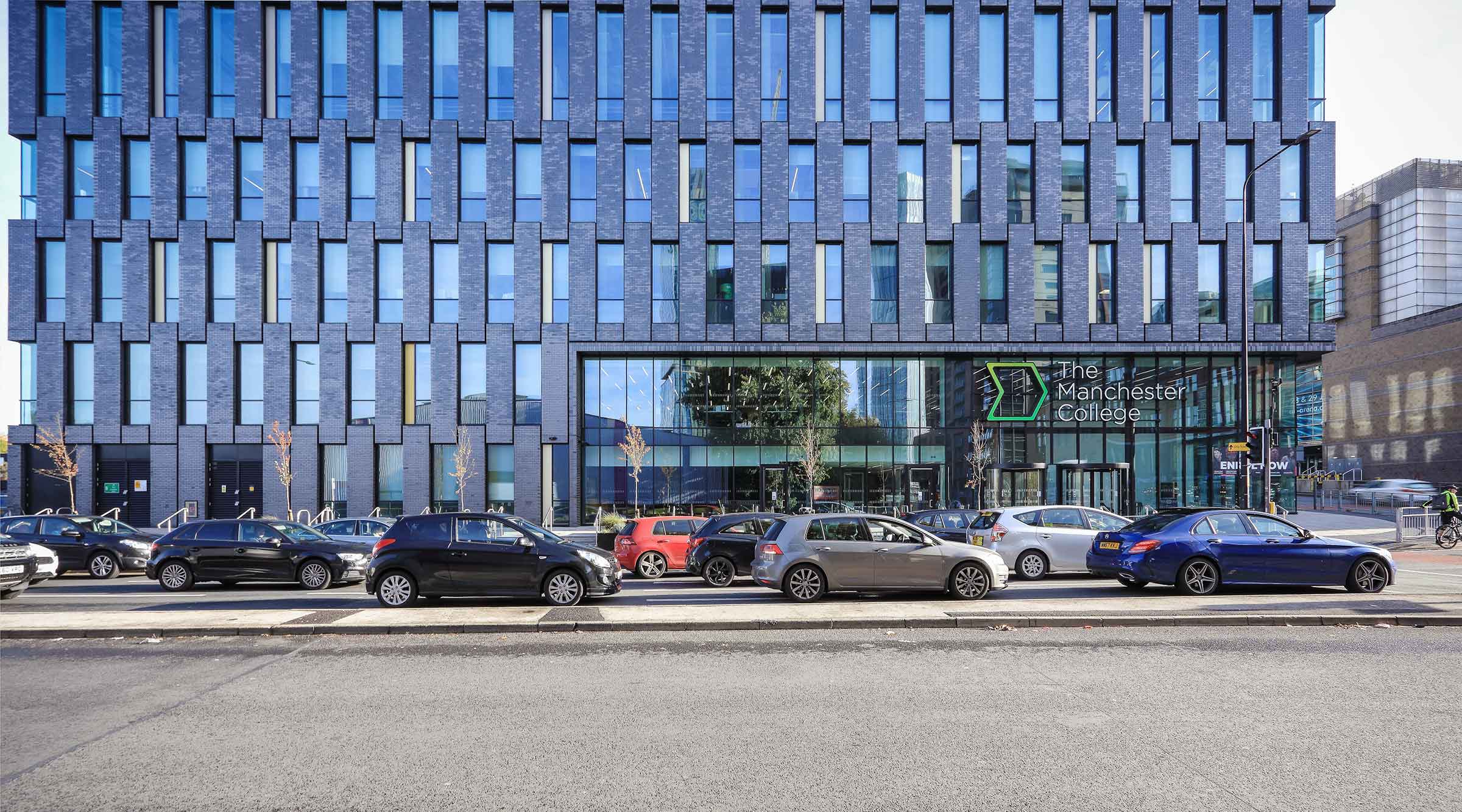 The side of a modern educational building. It is dark gray in colour and features rows of aluminium windows. There is a road in front of the building.