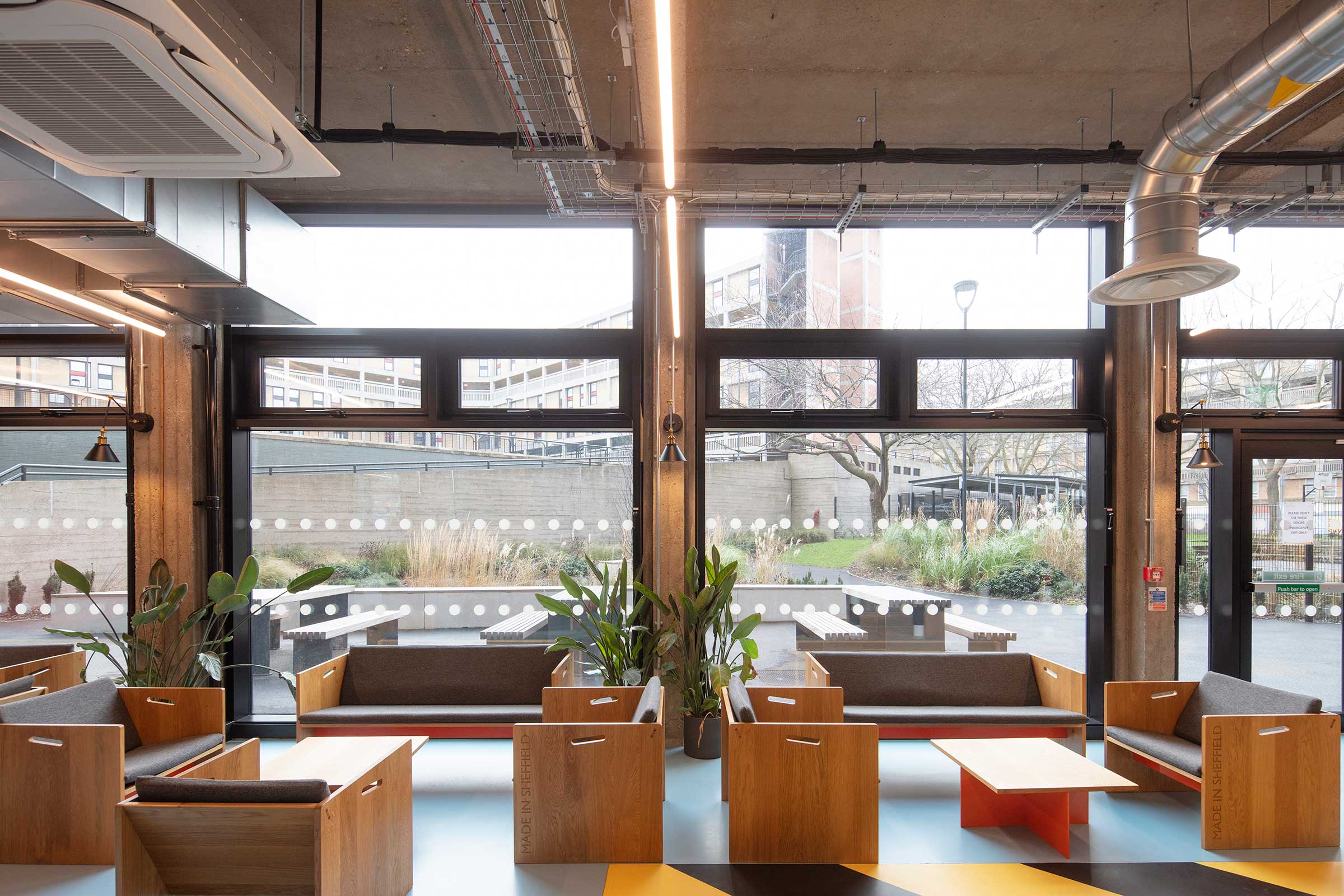 A large, well lit atrium with seating, tables and planting. There are dark gray windows to the rear of the building. 