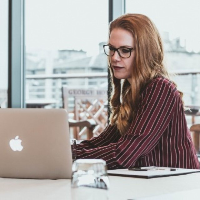 A woman with glasses is sitting at a computer and is using the software. She is wearing a red and black striped blazer.